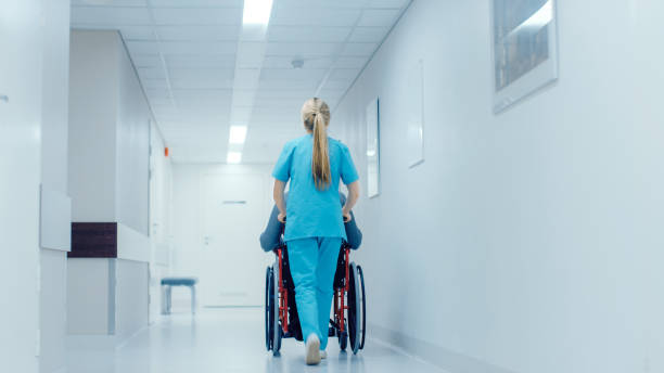 Shot of the Female Nurse Moving Patient in the Wheelchair Through the Hospital Corridor. Doing Procedures. Bright Modern Hospital with Friendly Staff. Shot of the Female Nurse Moving Patient in the Wheelchair Through the Hospital Corridor. Doing Procedures. Bright Modern Hospital with Friendly Staff. walking aide stock pictures, royalty-free photos & images