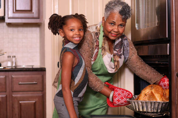 African descent family in home kitchen cooking Thanksgiving dinner. Family members work together in grandmother's home kitchen to prepare Thanksgiving dinner.  Grandmother takes roasted turkey out of oven with help from granddaughter. turkey meat photos stock pictures, royalty-free photos & images
