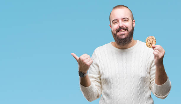 istock Young hipster man eating chocolate chips cookie over isolated background pointing and showing with thumb up to the side with happy face smiling 1043653410