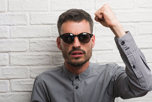 Young adult man wearing sunglasses standing over white brick wall annoyed and frustrated shouting with anger, crazy and yelling with raised hand, anger concept