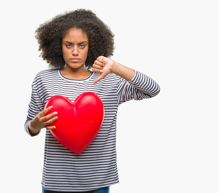 Young afro american woman holding red heart in love over isolated background with angry face, negative sign showing dislike with thumbs down, rejection concept