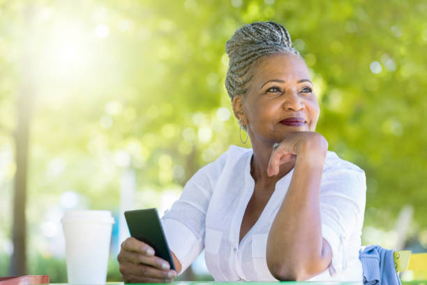 Senior woman waits for friend in public park A cheerful senior woman rests her chin in her hand as she looks away.  She is sitting at a table in a public park with coffee and her smart phone and waiting for a friend. black woman hair bun stock pictures, royalty-free photos & images