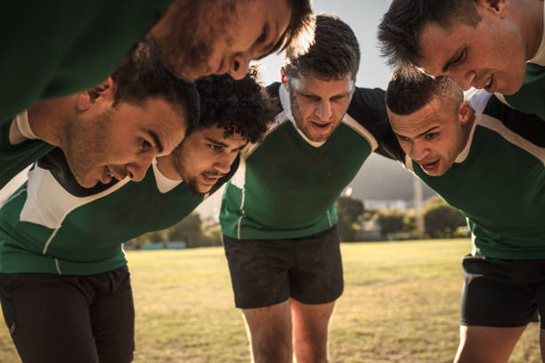 Group of young male rugby players in maroon jerseys compete for the loose ball on a sunny grassy field. The scene captures teamwork, energy, and sportsmanship with teammates observing.
