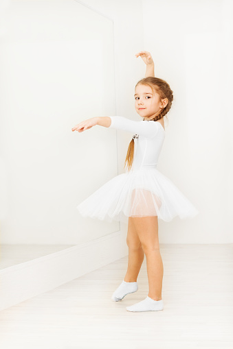 Adorable little girl in white dancewear, practicing posture during ballet class in light hall with copy-space