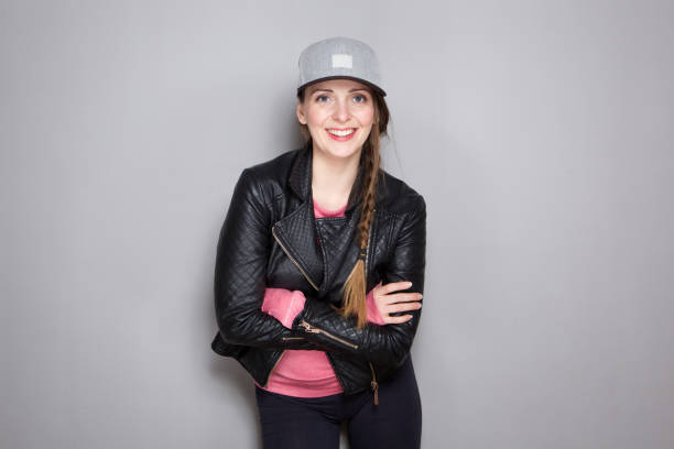 Portrait of cool young woman with leather jacket and hat standing by gray wall