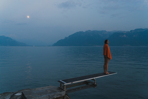 Young Caucasian woman in orange towel standing on diving board on Geneve lake at sunset