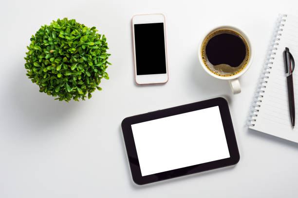Mock up of office desk with blank white screen tablet pc, blank black screen smartphone, coffee cup, notebook, pen and plant pot stock photo