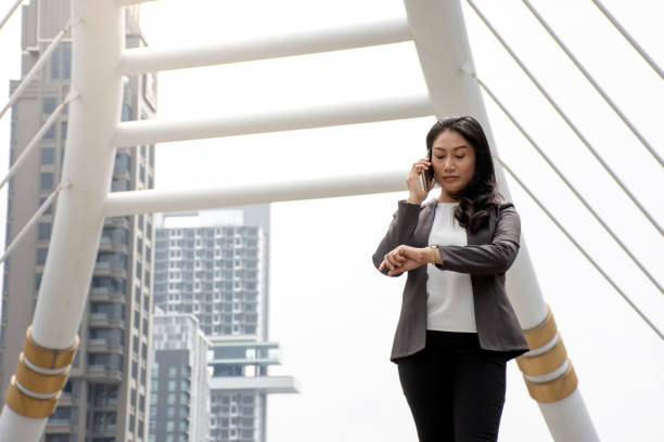 piękna azjatycka bizneswoman w formalnym garniturze rozmawiająca przez telefon patrząc na zegarek na rękę, czująca się zmęczona czekając na kogoś spóźniającego się - waiting women clock boredom zdjęcia i obrazy z banku zdjęć