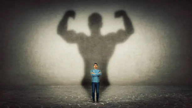 Confident businessman imagine himself a powerful hero, casting shadow of big strong muscular bodybuilder showing his biceps. Inner strength, leadership qualities. Business development.