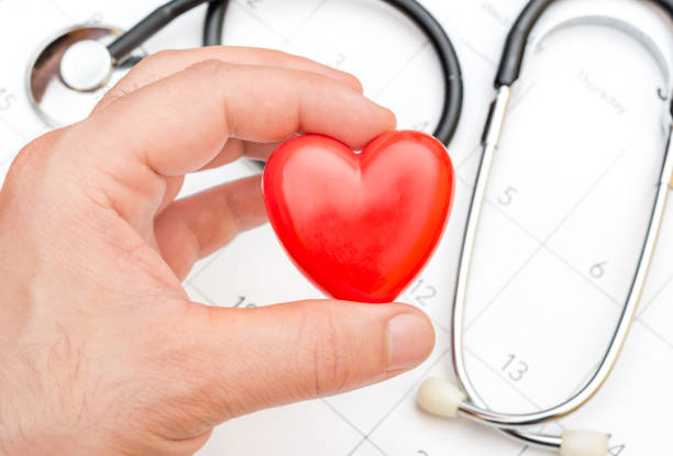 Man's hand holding red heart over calendar and stethoscope.
