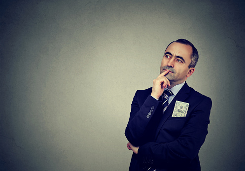 Adult businessman in formal suit holding finger on lips and looking away in contemplation on gray background