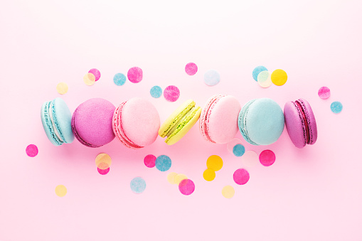 The row of colorful sweet macarons on pink background decorated with confetti. Top view, minimal styled.