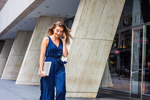 Young Eastern European American Woman talking on cell phone, traveling, working in New York City, wearing blue sleeveless jumpsuit, carrying laptop computer, walking on street outside office building.