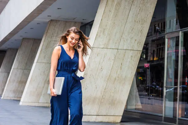 Young Eastern European American Woman talking on cell phone, traveling, working in New York City, wearing blue sleeveless jumpsuit, carrying laptop computer, walking on street outside office building."n