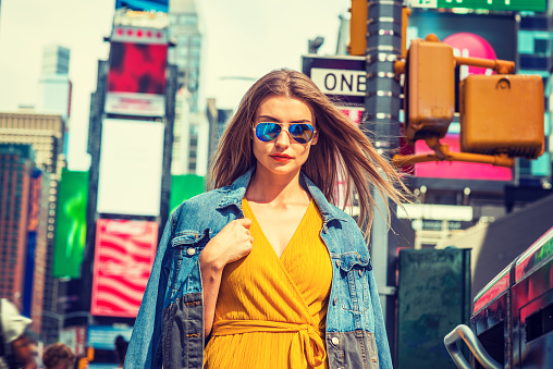 Young Eastern European Woman traveling in New York, with long brown hair, wearing yellow dress, blue Denim jacket draped over shoulder, blue sunglasses, standing on street in Times Square of Manhattan.