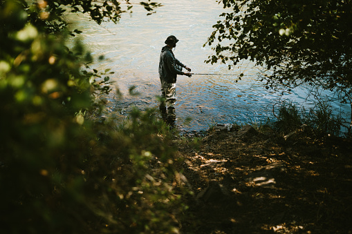 In silhouette of adult fisherman standing by the river and throwing the spinning. Lifestyle. Sport and hobby