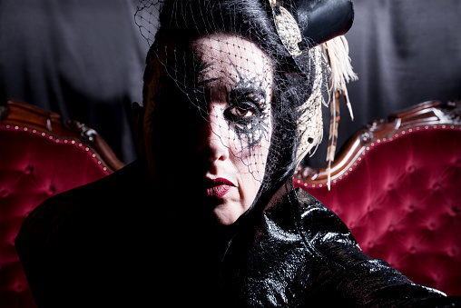 Scary goth styled lady in costume sitting on red tufted velvet sofa close-up looking at camera