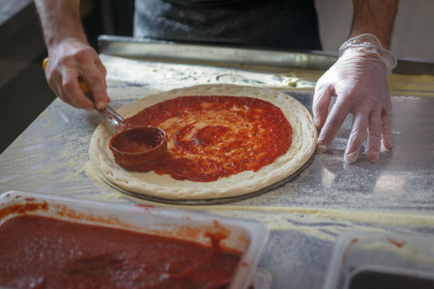 Baker spreads base of pizza with tomato sauce