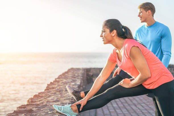 casal desportivo, esticando as pernas na frente do mar ao pôr do sol - jovens pessoas formação e fazendo as pernas exercícios ao ar livre - esporte, relacionamento, conceito de estilo de vida saudável - couple stretching running jogging - fotografias e filmes do acervo