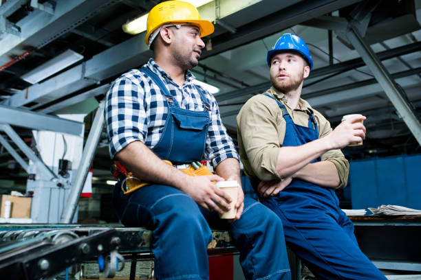 istock Content handsome multiethnic factory workers wearing hardhats and overalls talking at break and drinking coffee while sitting on conveyor belt 1043888364