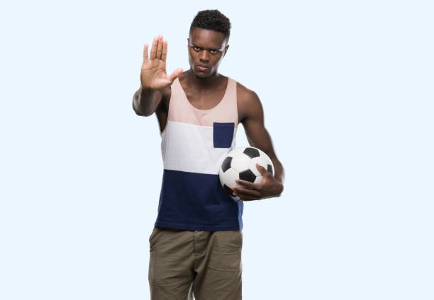 jovem americano africano, segurando a bola de futebol com a mão aberta, fazendo o sinal de stop, com expressão séria e confiante, gesto de defesa - football human hand holding american football - fotografias e filmes do acervo