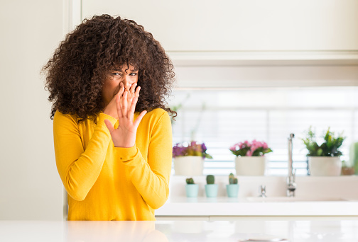 African american woman wearing yellow sweater at kitchen smelling something stinky and disgusting, intolerable smell, holding breath with fingers on nose. Bad smells concept.