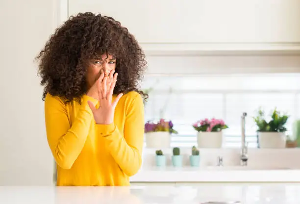 Photo of African american woman wearing yellow sweater at kitchen smelling something stinky and disgusting, intolerable smell, holding breath with fingers on nose. Bad smells concept.