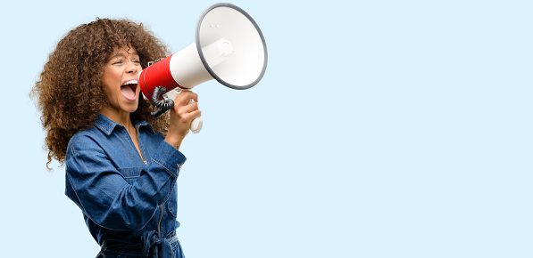 Mujer afroamericana que lleva mono azul comunica gritando fuerte sosteniendo un megáfono, expresando éxito y concepto positivo, idea marketing o ventas photo