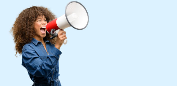 mujer afroamericana que lleva mono azul comunica gritando fuerte sosteniendo un megáfono, expresando éxito y concepto positivo, idea marketing o ventas - chillar fotografías e imágenes de stock