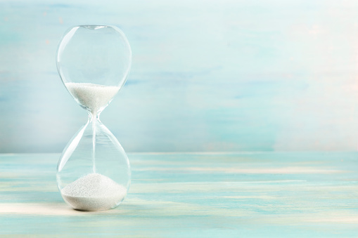 A side view of an hourglass with falling sand, on a teal background with copy space, slightly toned image