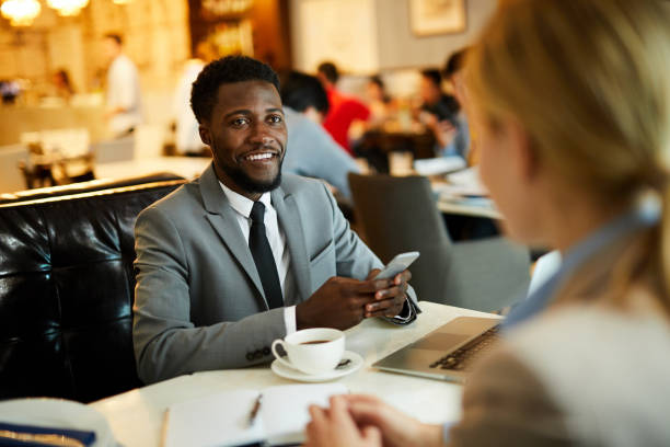 istock Cheerful optimistic young black businessman sitting at table and looking at colleague while using phone at informal meeting 1044046622