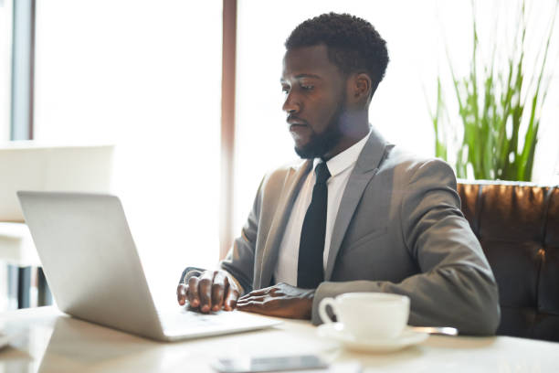 istock Serious busy black bearded man in gray jacket sitting at table and using laptop while working with files remotely 1044046772