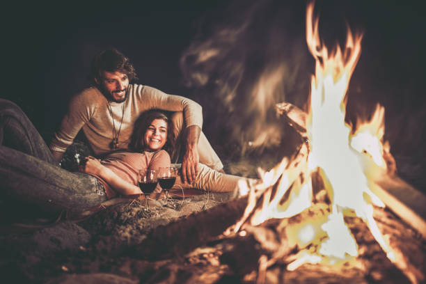 Happy relaxed couple enjoying in their date night by the campfire. Young happy couple relaxing on the beach by the campfire at night. Bonfire stock pictures, royalty-free photos & images