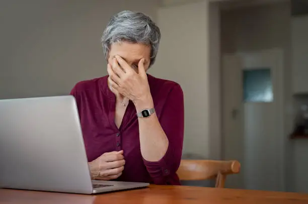 Mature businesswoman suffering a stress headache sitting at her desk with closed eyes in pain. Senior woman thinking about to complete work task. Depressed tired mature lady suffering from chronic daily headache from computer.