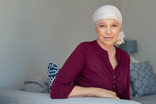 Mature woman with cancer in pink headscarf smiling sitting on couch at home. Smiling woman suffering from cancer sitting after taking chemotherapy sessions. Portrait of mature lady facing side-effects of hair loss, copy space.