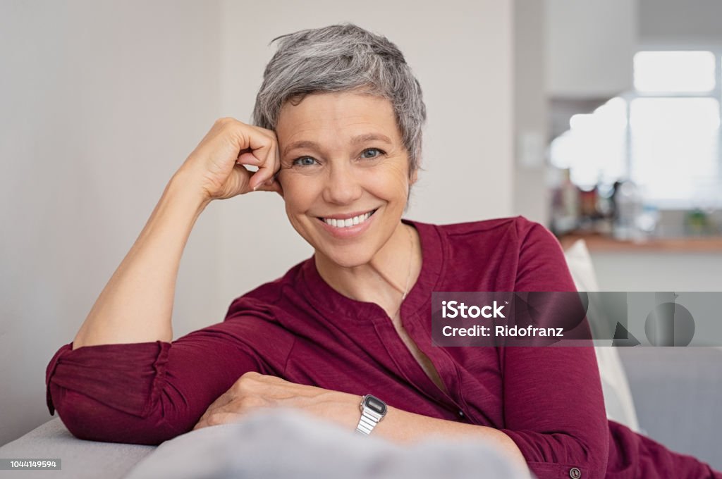 Happy senior woman on couch Portrait of smiling senior woman relaxing on couch at home. Happy mature woman sitting on sofa and looking at camera. Closeup of lady relaxing at home. Women Stock Photo