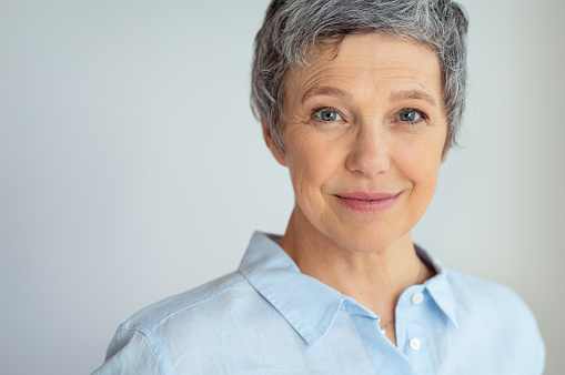 Closeup face of senior business woman standing against grey background with copy space. Portrait of successful woman in blue shirt feeling confident and looking at camera. Happy mature woman face standing.
