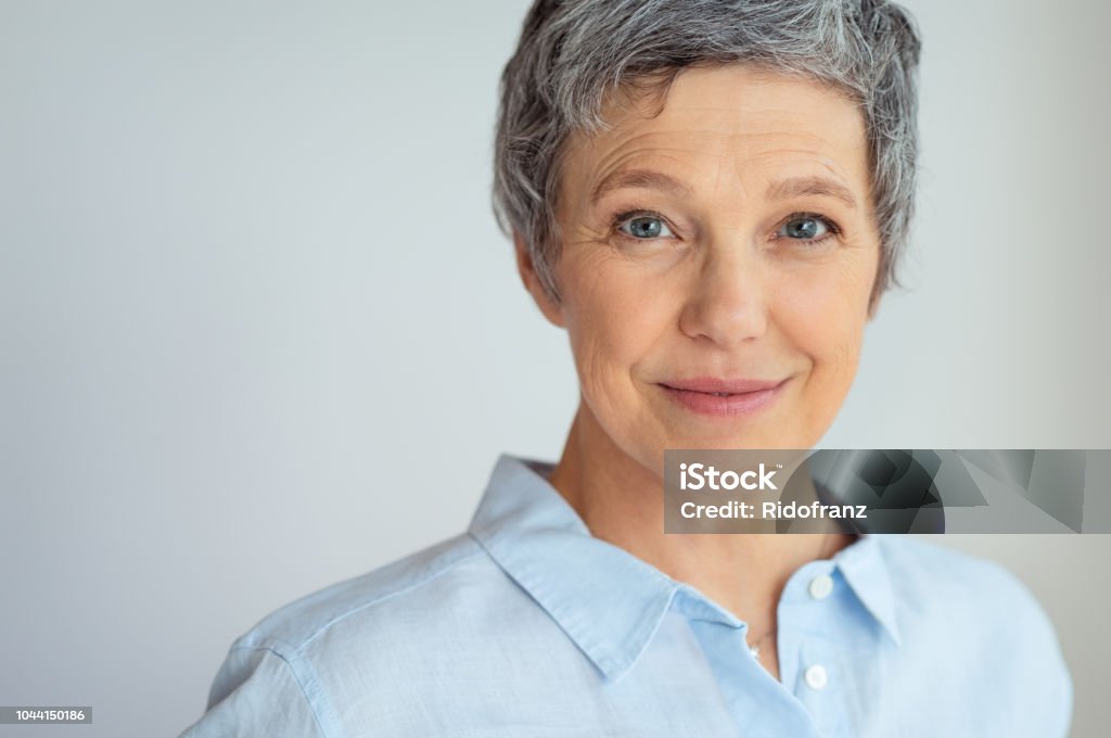 Smiling senior woman Closeup face of senior business woman standing against grey background with copy space. Portrait of successful woman in blue shirt feeling confident and looking at camera. Happy mature woman face standing. Women Stock Photo