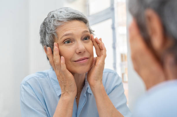 Mature woman aging Beautiful senior woman checking her face skin and looking for blemishes. Portrait of mature woman massaging her face while checking wrinkled eyes in the mirror. Wrinkled lady with grey hair checking wrinkles around eyes, aging process. mirror stock pictures, royalty-free photos & images
