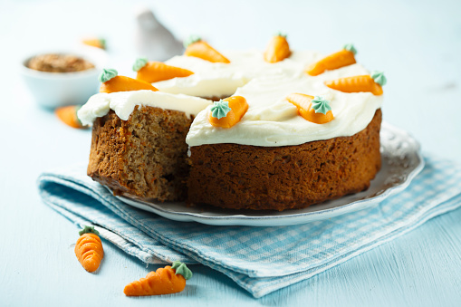 Stock photo showing close-up view of single-use cardboard container with carrot cake slice on black paper serviette. The carrot cake is topped with a rich cream cheese icing and decorated with an orange fondant carrot decoration.