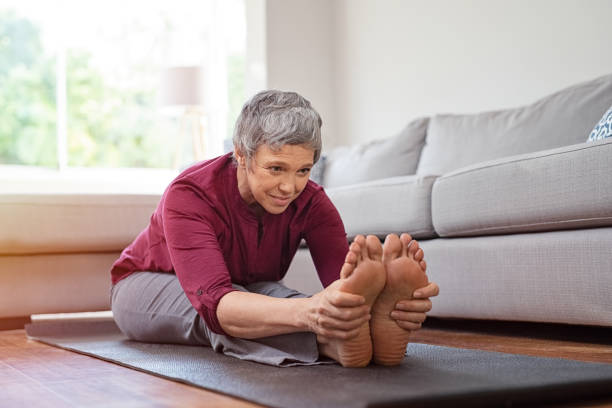 Mature woman doing yoga exercise at home Beautiful senior woman doing stretching exercise while sitting on yoga mat at home. Mature woman exercising in sportswear by stretching forward to touch toes. Healthy active lady doing yoga and flexibility exercise. mature woman barefoot stock pictures, royalty-free photos & images