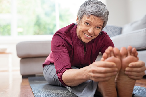 Closeup of senior woman stretching to touch toes while sitting on yoga mat. Portrait of mature woman doing her stretches at home while looking at camera. Happy healhty lady doing yoga exercises.