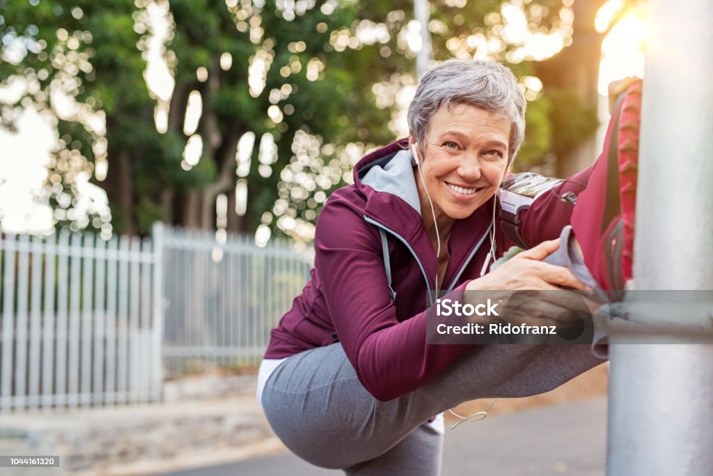 Mature woman warming up before jogging Smiling retired woman listening to music while stretching legs outdoors. Senior woman enjoying daily routine warming up before running at morning. Sporty lady doing leg stretches before workout and looking at camera. Exercising Stock Photo