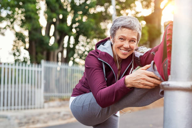Mature woman warming up before jogging Smiling retired woman listening to music while stretching legs outdoors. Senior woman enjoying daily routine warming up before running at morning. Sporty lady doing leg stretches before workout and looking at camera. DisruptAgingCollection stock pictures, royalty-free photos & images