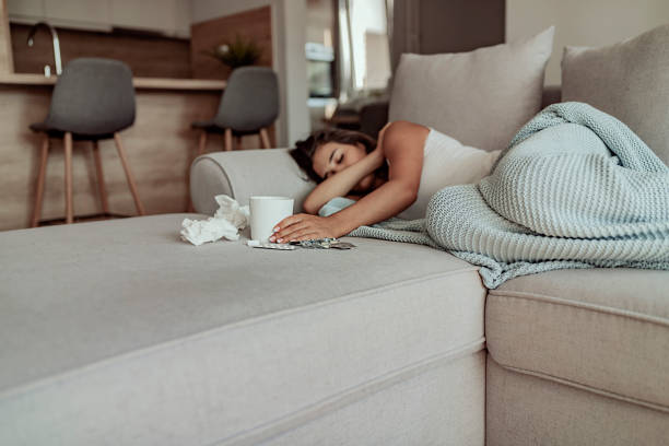Resting to get better Sick Woman Sleeps Covered With a Blanket Lying in Bed With High Fever and a Flu, Resting at Living Room. She Is Exhausted and Suffering From Flu With Cup of Tea, Medicaments and Tissues sofa bed stock pictures, royalty-free photos & images
