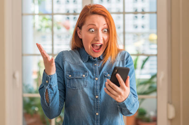 istock Redhead woman using smartphone at home very happy and excited, winner expression celebrating victory screaming with big smile and raised hands 1044224148