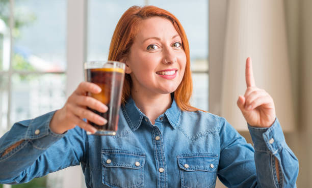 istock Redhead woman holding soda refreshment surprised with an idea or question pointing finger with happy face, number one 1044224334
