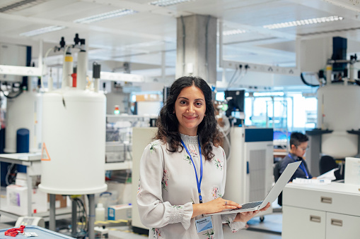 A portrait of a mixed race female scientist.She stands and looks confidently into the camera, her medical laboratory, is full of specialist, state of the art equipment. This this is a first class research facility, can be used to detect, cancer, diabetes, autism and dementia. A coworker is out of focus in the background. This pleasant woman is looking up from her personal computer.