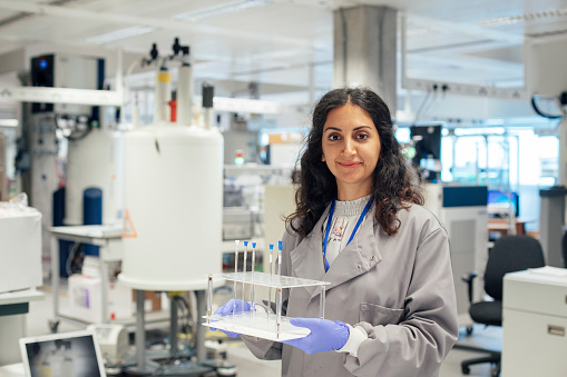 A mixed race female scientist stands in her laboratory, she is holding body fluid samples and is wearing protective workwear. In the background there is a vast array of specialist research equipment, she is about to run some human medical samples. New forms of innovation are being trialed, with investment in new technologies.