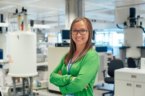 A female science graduate stands in her working laboratory, she has a confident smile, and looks directly into camera.The area behind her, is full of specialist disease detection equipment, including NMR machines. A role model to the next generation.